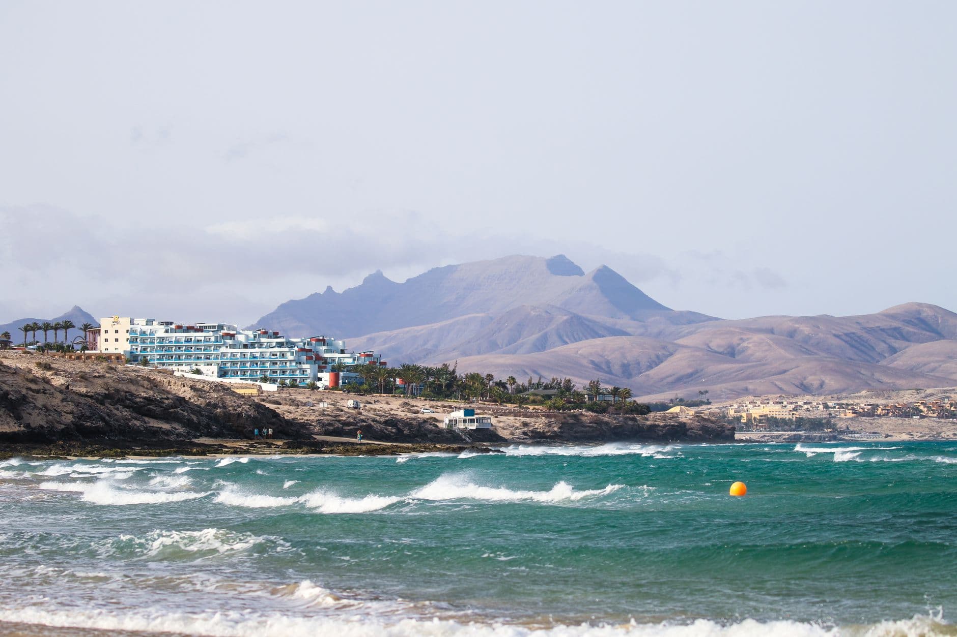 De kust bij Costa Calma op Fuerteventura, met golven op de voorgrond, een hotel op de rotskust en bergen op de achtergrond