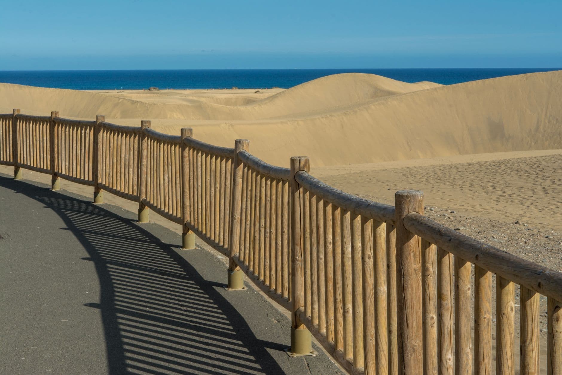 Een geasfalteerd wandelpad met een houten hek langs de zandduinen van Maspalomas op Gran Canaria, met de zee in de verte