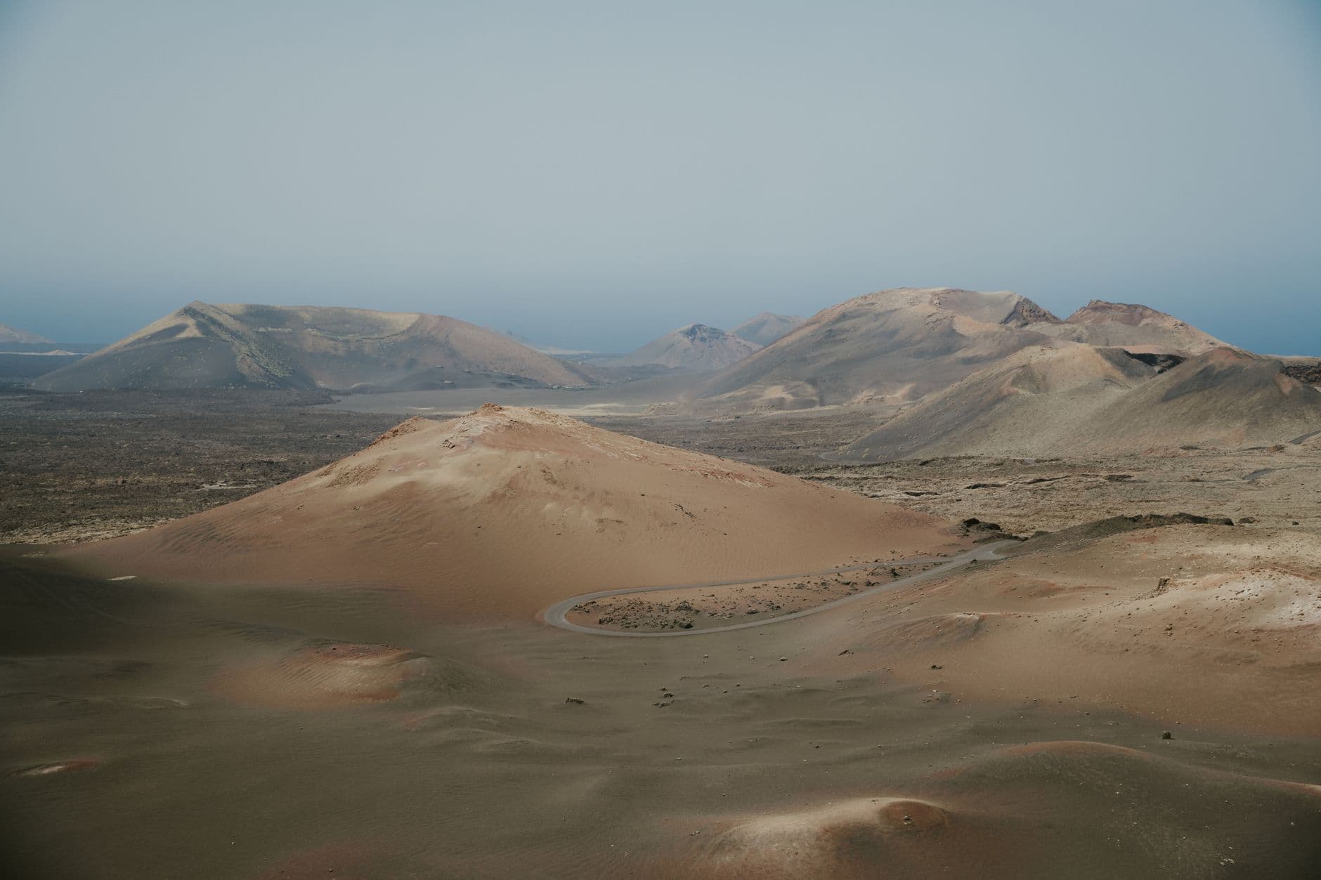 Vulkaankegels en een kronkelende weg door een kaal vulkanisch landschap op Lanzarote, onder een heiige lucht