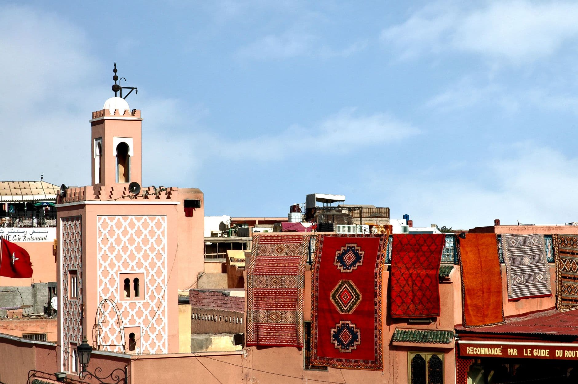 Kleurrijke tapijten hangen over een dakrand in Marrakech, met links een roze minaret