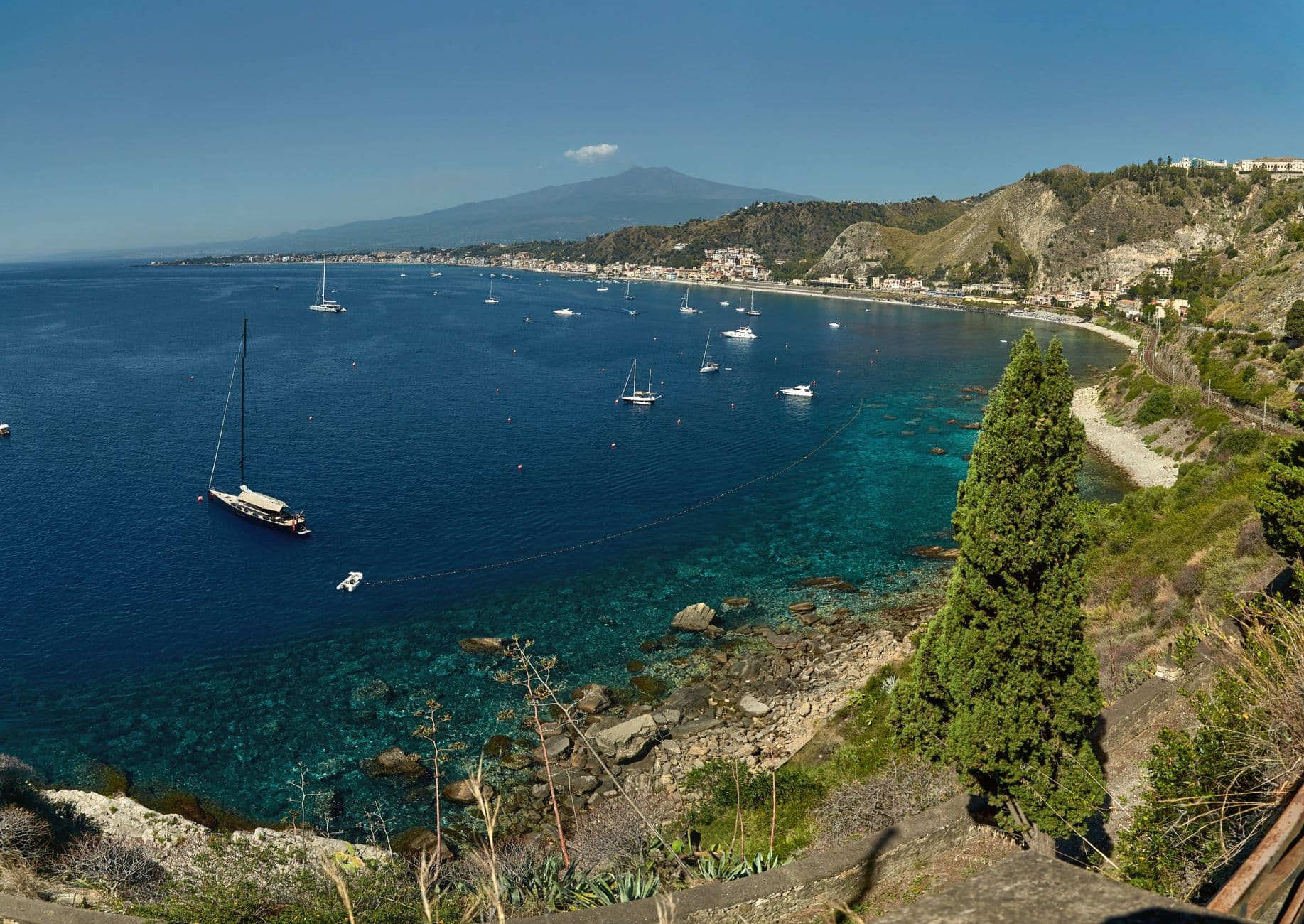 Stunning view of the Mediterranean coast featuring anchored sailboats and a distant mountain range under a clear blue sky.