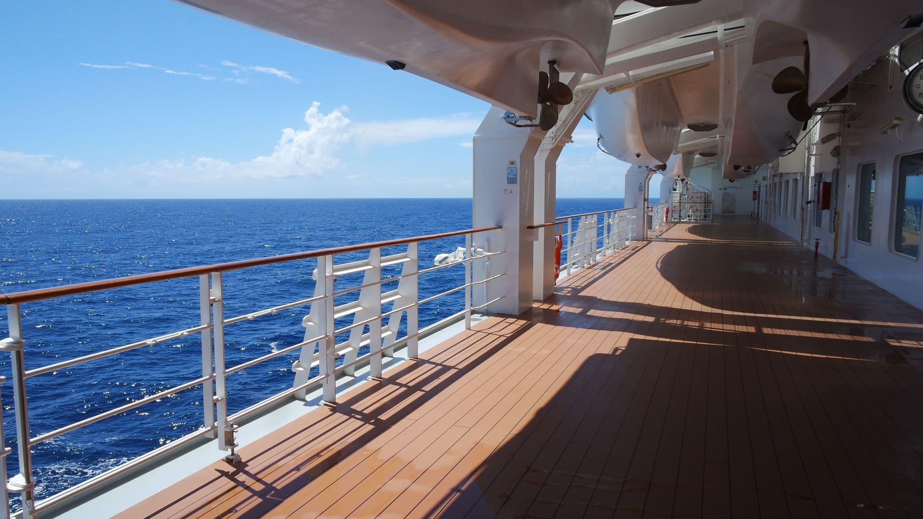 Spacious cruise ship deck with lifeboats, railings, and open ocean view under clear blue sky.