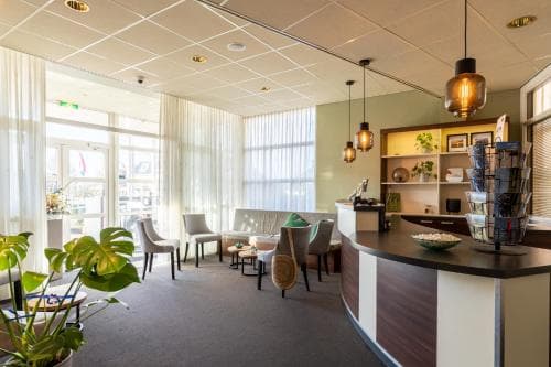 Ruime hotellobby met receptiebalie, zithoek met grijze stoelen en bank, en grote ramen met wit gordijnen