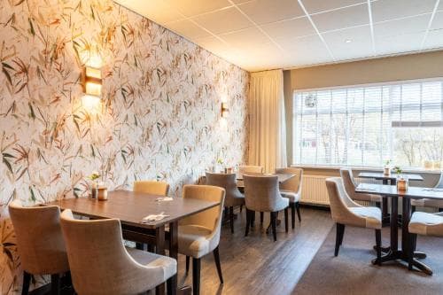 Leeg restaurant met beige stoelen, houten tafels, bloemenbehang en grote ramen.