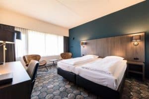 Hotelkamer met twee opgemaakte eenpersoonsbedden, een bureau, twee leren stoelen, en een groot raam. De muur achter het bed is donkerblauw
