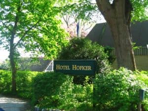 Groen bord met witte letters "HOTEL HOFKER" tussen weelderig groen gebladerte en bomen. Op de achtergrond is een dak van een gebouw zichtbaar.