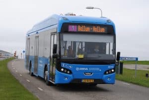 Blauwe Arriva bus met bestemming "1 Ballum-Hollum" rijdt op een weg met witte stippellijnen en gras aan weerszijden. De bus heeft nummer "3019" en kenteken "23-BHX-5". Op de achtergrond een bewolkte lucht, gebouw, verkeersbord en grasveld.