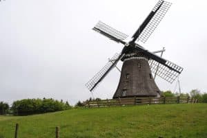 Grote, donkere molen met wieken op een heuvel met groen gras, omringd door een houten hek en bomen op de achtergrond. De lucht is grijs.