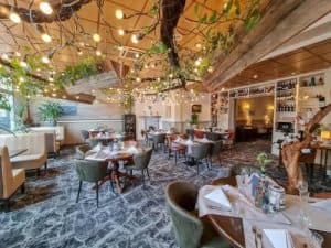 Interieur van een restaurant met houten elementen, planten tegen het plafond met lampjes ertussen, gedekte tafels met stoelen en glazen wanden met zichtbare flessen achterin de zaal.