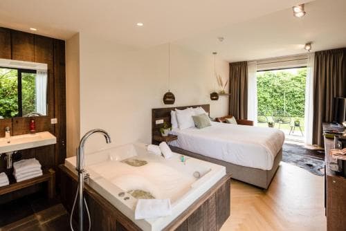 Hotelkamer met een wit opgemaakt bed, een grote jacuzzi in het midden van de kamer, een wastafel met handdoeken, en openslaande deuren naar een terras met groen.
